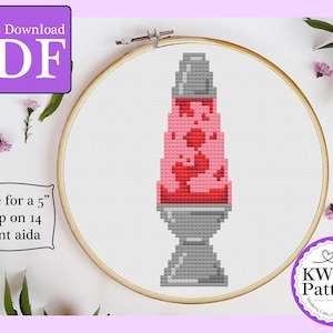 May include: Cross-stitch design of a lava lamp in an embroidery hoop. The lamp is pink and red, with a grey base. The pattern is for a 12.7 cm hoop on 14-count aida fabric. The image includes the text "Instant Download PDF".