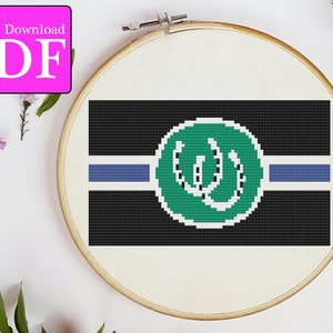 May include: A cross-stitch pattern of a green circle with a white 'W' inside, on a black background with a blue stripe. The pattern is in a wooden embroidery hoop.