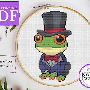 Puede incluir: Patrón de punto de cruz de una rana verde que lleva un sombrero de copa negro con una banda roja y un esmoquin morado. El patrón cabe en un aro de 15 cm en tela Aida de 18 puntos. KW Patterns - Artesanía moderna, punto de cruz único.