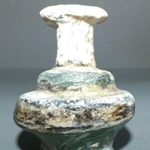 Puede incluir: Una pequeña botella de vidrio antigua con una parte superior blanca y un cuerpo bulboso. La botella tiene una combinación de colores verde y marrón, con una apariencia desgastada. La forma de la botella recuerda a los recipientes antiguos.