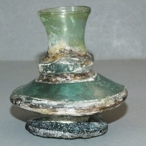 Puede incluir: Una pequeña botella de vidrio verde antiguo con una base acampanada y un cuello estrecho y acanalado. La botella tiene un aspecto envejecido con una banda oscura y texturizada en el medio. La base mide unos 4 cm de diámetro.