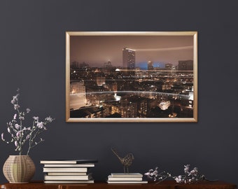 May include: A framed photograph of a city skyline at night. The city is lit up with lights, and there are tall buildings in the distance. The photo is in a gold frame and is hanging on a dark wall.