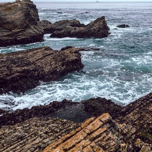 May include: A rocky coastline with waves crashing against the shore. The rocks are dark brown and grey, and the water is a deep blue.