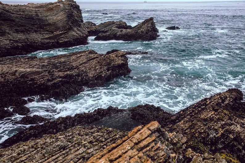 May include: A rocky coastline with waves crashing against the shore. The rocks are dark brown and grey, and the water is a deep blue.