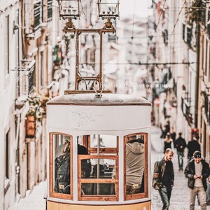 Puede incluir: Un tranv&iacute;a amarillo vintage viaja por una calle adoquinada en una ciudad. El tranv&iacute;a tiene un techo blanco y ventanas de madera. El tranv&iacute;a est&aacute; subiendo una colina empinada. Hay gente caminando por la calle detr&aacute;s del tranv&iacute;a.