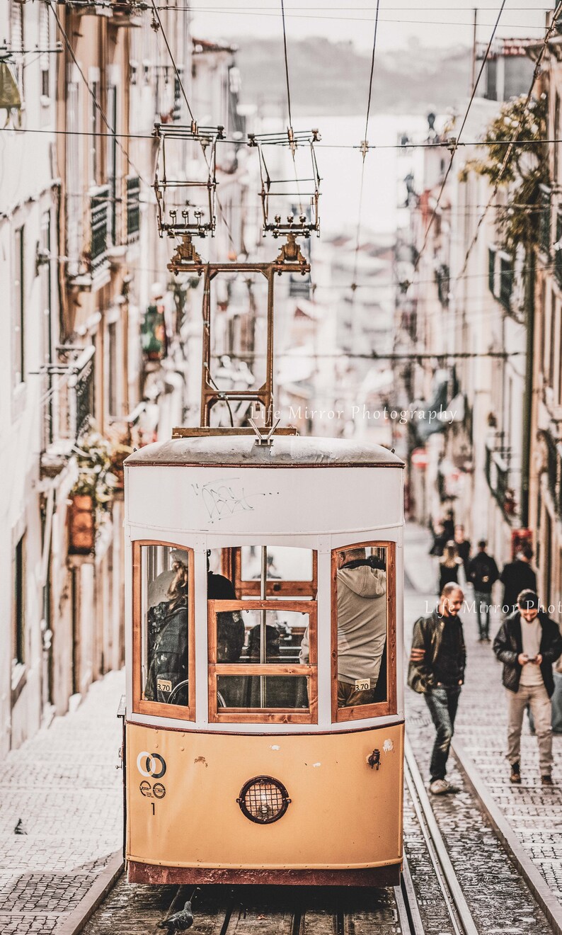 Puede incluir: Un tranv&iacute;a amarillo vintage viaja por una calle adoquinada en una ciudad. El tranv&iacute;a tiene un techo blanco y ventanas de madera. El tranv&iacute;a est&aacute; subiendo una colina empinada. Hay gente caminando por la calle detr&aacute;s del tranv&iacute;a.