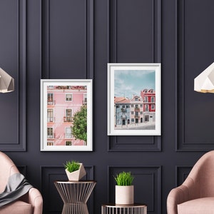 Op de afbeelding: Twee ingelijste prints van kleurrijke gebouwen in een stad, hangend aan een donkergrijze muur met witte lijstwerk. Twee witte papieren hanglampen hangen boven twee roze fauteuils.