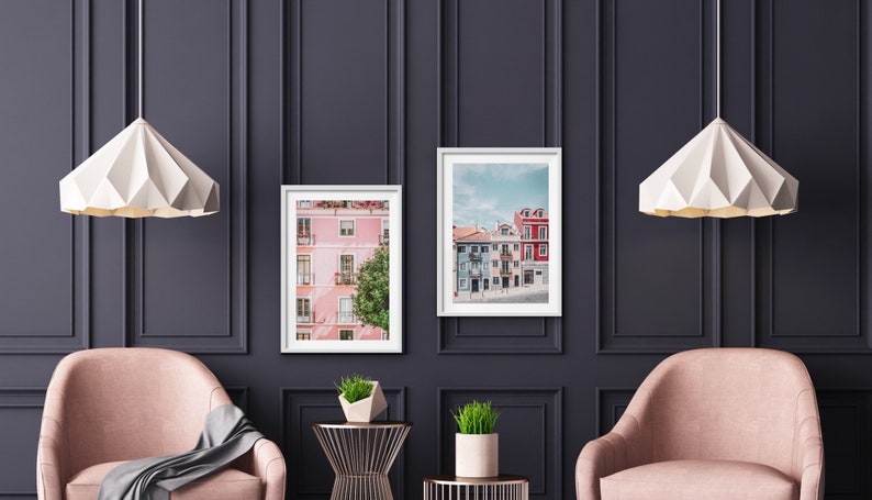 Op de afbeelding: Twee ingelijste prints van kleurrijke gebouwen in een stad, hangend aan een donkergrijze muur met witte lijstwerk. Twee witte papieren hanglampen hangen boven twee roze fauteuils.