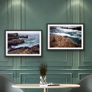 May include: Two framed photographs of ocean waves crashing against rocky shores. The photos are hung on a green wall with gold wall sconces on either side.