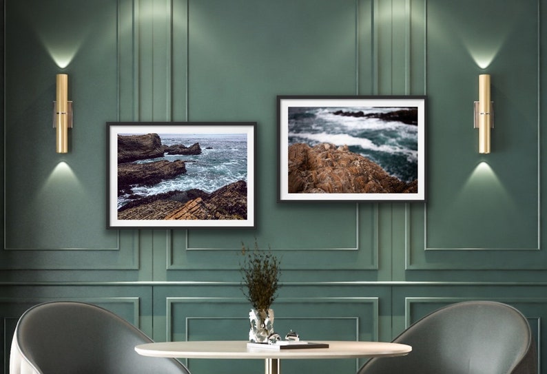 May include: Two framed photographs of ocean waves crashing against rocky shores. The photos are hung on a green wall with gold wall sconces on either side.