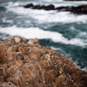 May include: Close-up of rugged, brown rock formations with a blurred background of blue ocean water and white foam.