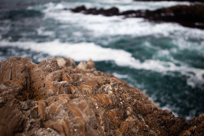 May include: Close-up of rugged, brown rock formations with a blurred background of blue ocean water and white foam.