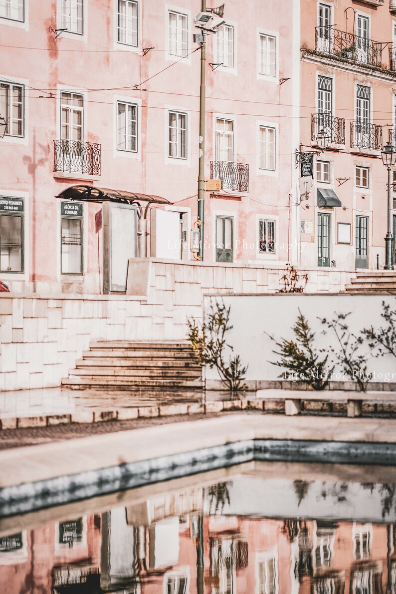 Puede incluir: Un reflejo de un edificio rosa con ventanas y balcones en una peque&ntilde;a piscina de agua. El edificio se encuentra en un entorno urbano con una pared de ladrillo blanco y escalones en primer plano.