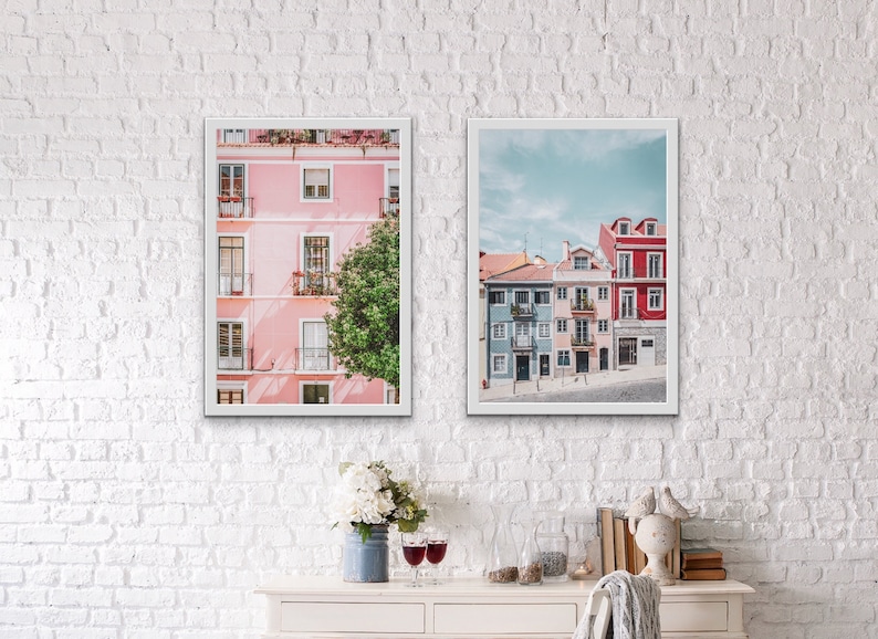 Op de afbeelding: Twee ingelijste foto's van kleurrijke gebouwen in Lissabon, Portugal. De linker foto toont een roze gebouw met witte versieringen en een groene boom. De rechter foto toont een rij blauwe, roze en rode gebouwen met witte versieringen.