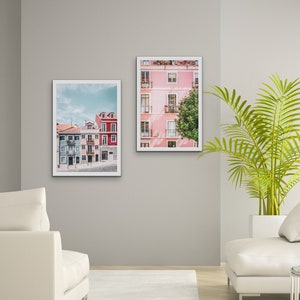 Op de afbeelding: Twee ingelijste foto's van kleurrijke gebouwen in een stad. De foto links toont een rij gebouwen met blauwe, groene en rode gevels. De foto rechts toont een roze gebouw met witte versieringen en een boom ervoor.