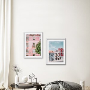 Op de afbeelding: Twee ingelijste foto's van kleurrijke gebouwen in een stad. De eerste foto toont een roze gebouw met witte versieringen en een groene boom ervoor. De tweede foto toont een rij kleurrijke gebouwen met blauwe, roze en rode gevels.