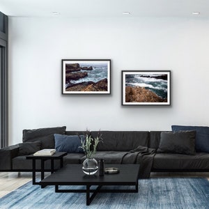May include: Two framed photographs of ocean waves crashing against rocky shores. The photos are hung on a white wall above a black leather sofa in a modern living room.