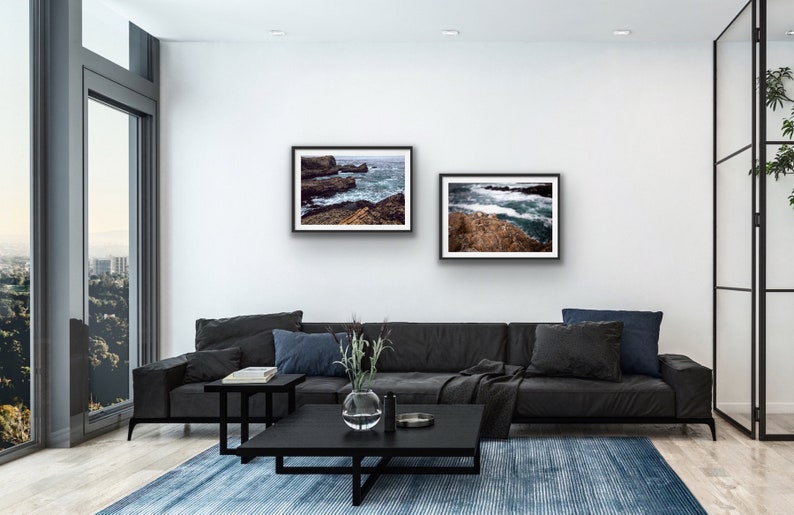 May include: Two framed photographs of ocean waves crashing against rocky shores. The photos are hung on a white wall above a black leather sofa in a modern living room.