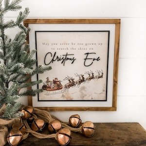May include: A framed Christmas Eve sign with a vintage illustration of Santa's sleigh and reindeer. The sign reads "May you never be too grown up to search the skies on Christmas Eve." Copper bells and a small Christmas tree are on a wooden shelf.