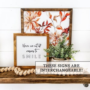 May include: Two framed signs with interchangeable designs. The top sign features a watercolor floral print with orange, red, and green leaves. The bottom sign has a white background with black text that reads "Never run out of reasons to SMILE".