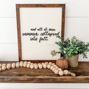 May include: A wooden sign with the quote "and all at once, summer collapsed into fall." by Oscar Wilde. The sign is framed in brown wood and has a small wooden leaf attached to the bottom right corner. The sign is sitting on a wooden shelf with a string of wooden beads and a small coconut-like object.