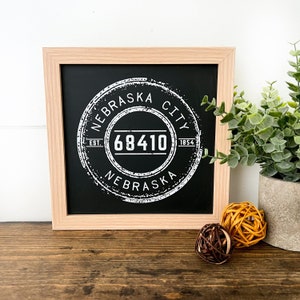 May include: A black framed chalkboard sign with a white distressed circle design. The circle says "Nebraska City Est. 1854" and has the number "68410" in the centre. The sign is sitting on a wooden surface.