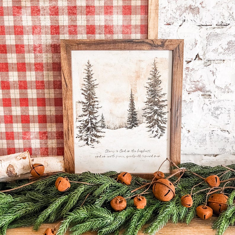 May include: A framed print with a wintery forest scene and the text "Glory to God in the highest, and on earth peace, goodwill toward men." The print is surrounded by a garland of evergreen branches with brown jingle bells.
