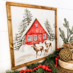 May include: A watercolor painting of a red cabin in the woods with snow on the ground. Two deer are walking in front of the cabin. The painting is framed in a brown wood frame.