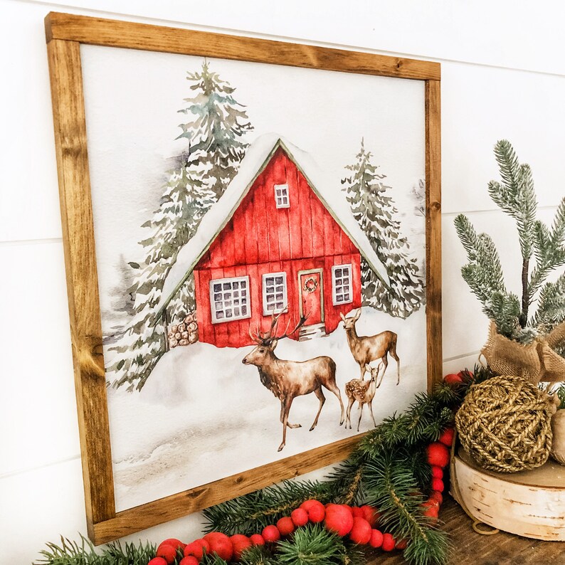 May include: A watercolor painting of a red cabin in the woods with snow on the ground. Two deer are walking in front of the cabin. The painting is framed in a brown wood frame.
