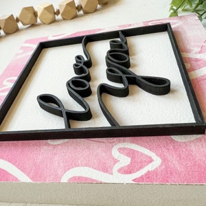 May include: A black wooden sign with the words "Love You" in a cursive font. The sign is framed with a black border and is sitting on a pink background with white hearts.