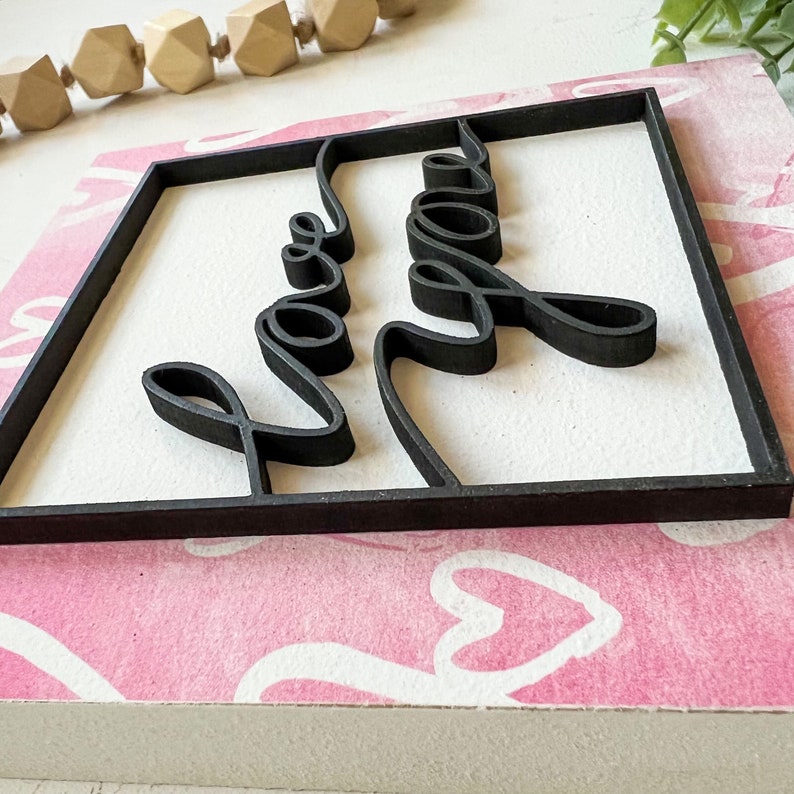 May include: A black wooden sign with the words "Love You" in a cursive font. The sign is framed with a black border and is sitting on a pink background with white hearts.