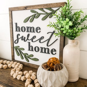 Puede incluir: Un letrero de madera con el texto "home sweet home" en letras negras. El letrero est&aacute; decorado con hojas verdes y se exhibe en un estante con un jarr&oacute;n de vegetaci&oacute;n y un cuenco decorativo.