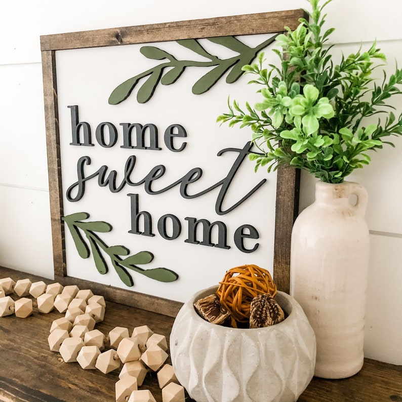 Puede incluir: Un letrero de madera con el texto "home sweet home" en letras negras. El letrero est&aacute; decorado con hojas verdes y se exhibe en un estante con un jarr&oacute;n de vegetaci&oacute;n y un cuenco decorativo.