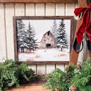 May include: A framed print shows a winter scene with a barn and snow-covered trees. The artwork is in a dark brown wooden frame. The scene includes deer in the snow. The image is decorated with evergreen branches and a red velvet bow.