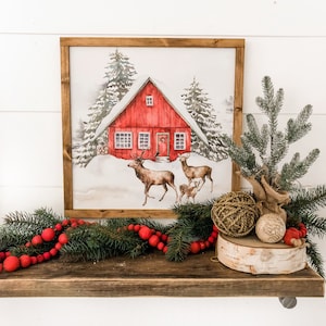 May include: A framed watercolor painting of a red cabin in a snowy forest with two deer and a fawn in the foreground. The cabin has a wreath on the door and snow on the roof. The painting is on a wooden shelf with a green garland and red berries.