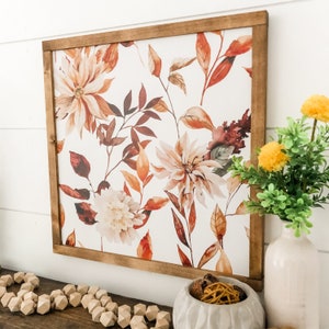 May include: A framed watercolor print with a floral design featuring brown, orange, and white flowers on a white background. The frame is made of wood and has a brown stain.