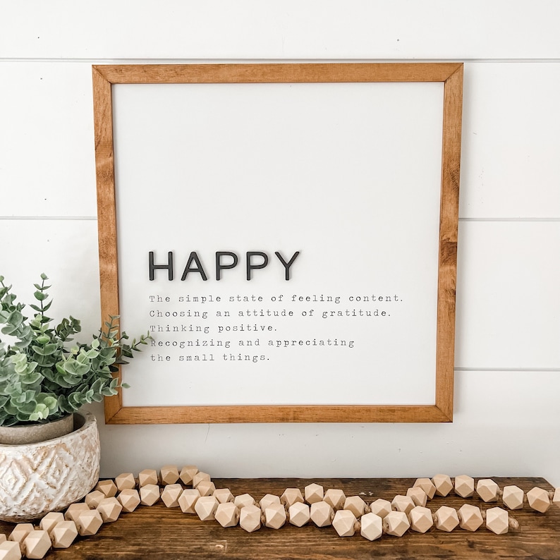 May include: A framed print with the word "HAPPY" in large black letters and a definition of happiness in smaller black text. The definition reads: "The simple state of feeling content. Choosing an attitude of gratitude. Thinking positive. Recognizing and appreciating the small things."