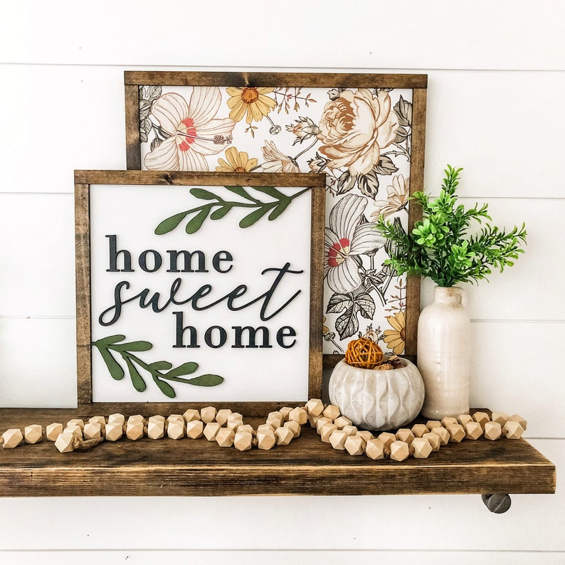 Puede incluir: Un letrero de madera con el texto "home sweet home" en letras negras y hojas verdes. El letrero est&aacute; en un estante con un letrero de patr&oacute;n floral detr&aacute;s, un peque&ntilde;o jarr&oacute;n con plantas verdes y un taz&oacute;n decorativo con una peque&ntilde;a bola naranja dentro.