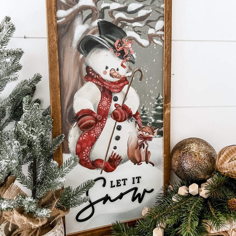 May include: A vintage-style framed print featuring a snowman with a red scarf and a fox. The snowman is holding a stick and the fox is sitting next to him. The print has the text "Let it Snow" in black cursive lettering.