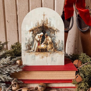 May include: An arched painting of the Nativity scene, depicting Mary, Joseph, and baby Jesus in a manger, with a cross atop a stable. The painting is set on a stack of red-covered books, with decorative bells and greenery.