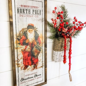 May include: A vintage-style wooden sign with a red and white illustration of Santa Claus delivering gifts. The sign reads "Reindeer Express, North Pole Delivery Co. Spreading Christmas Cheer Toys Games Gifts".