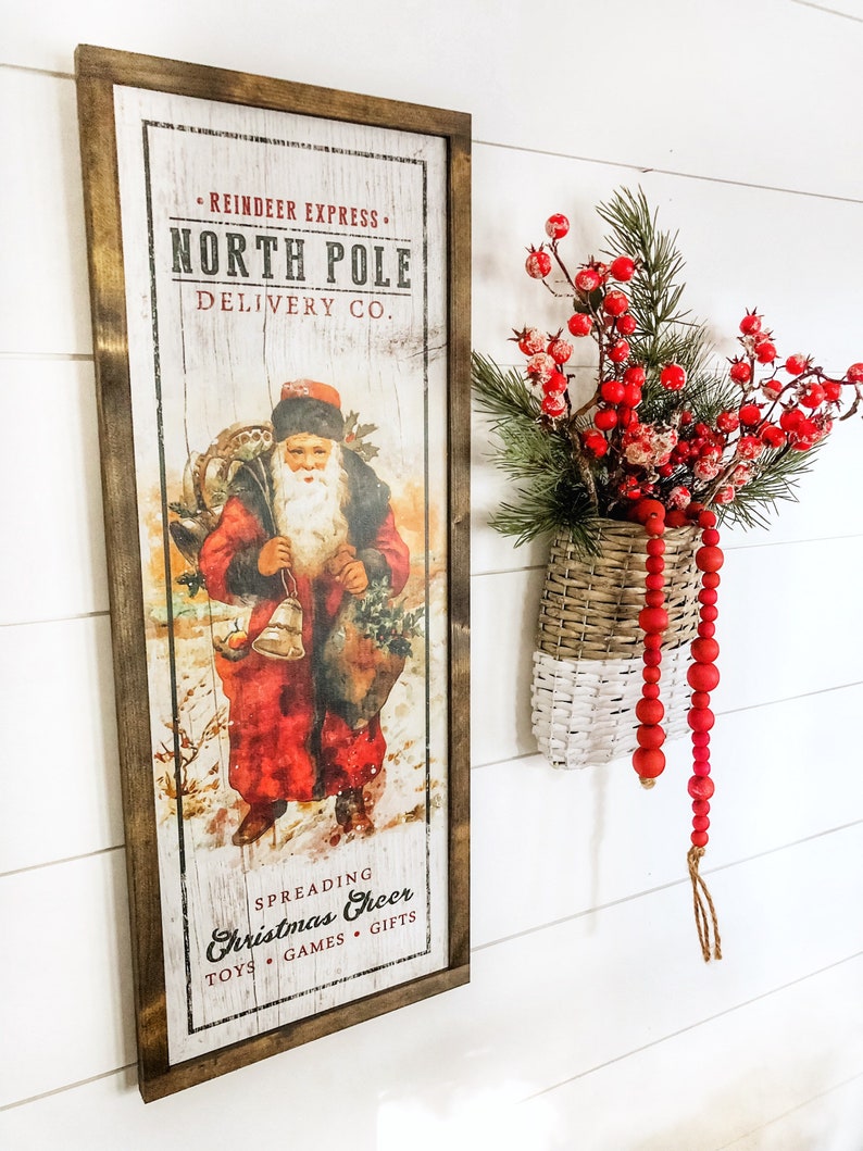May include: A vintage-style wooden sign with a red and white illustration of Santa Claus delivering gifts. The sign reads "Reindeer Express, North Pole Delivery Co. Spreading Christmas Cheer Toys Games Gifts".