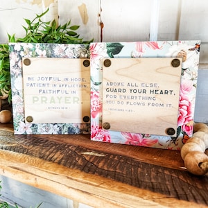 May include: Two wooden signs with floral designs and inspirational quotes. The first sign says "Be Joyful in Hope, Patient in Affliction, Faithful in Prayer." The second sign says "Above all else, Guard your heart, for everything you do flows from it."