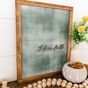 May include: A framed green sign with the text "but I think I love fall most of all..." in white lettering. The sign is hanging on a white wall with a wooden frame.