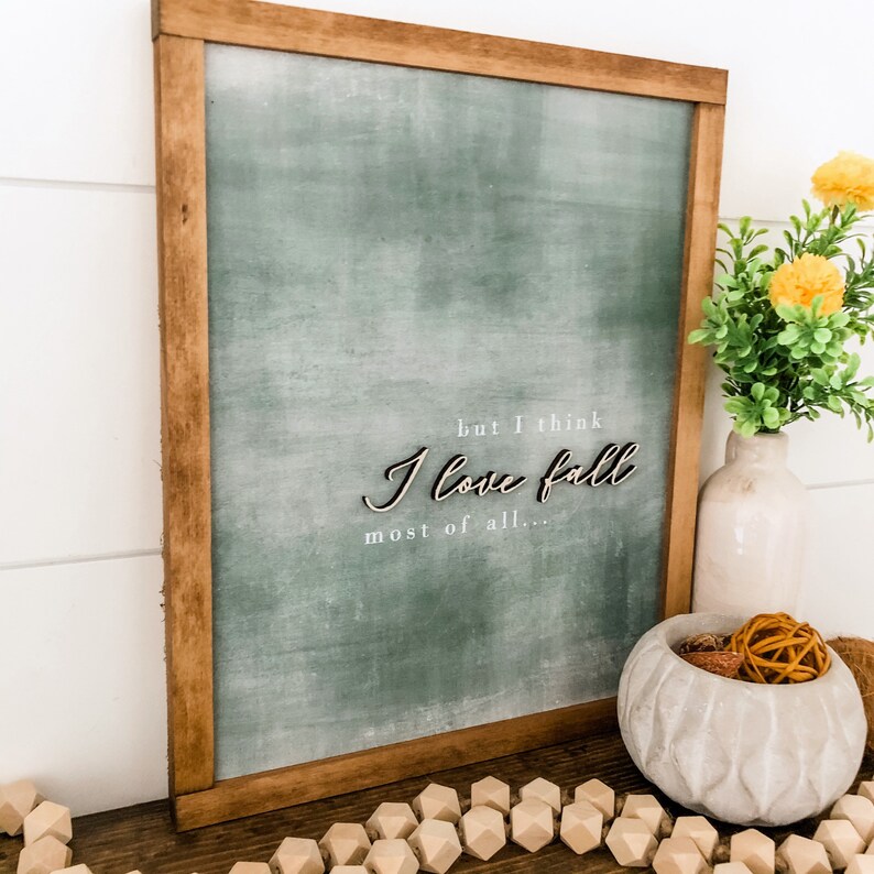May include: A framed green sign with the text "but I think I love fall most of all..." in white lettering. The sign is hanging on a white wall with a wooden frame.