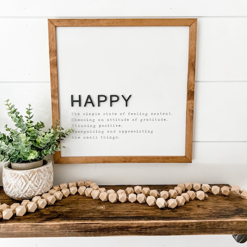 May include: A framed print with the word "HAPPY" in large black letters. The definition of happy is written in black text below the word. The print is hanging on a white wall above a wooden shelf with a small potted plant and a wooden bead garland.