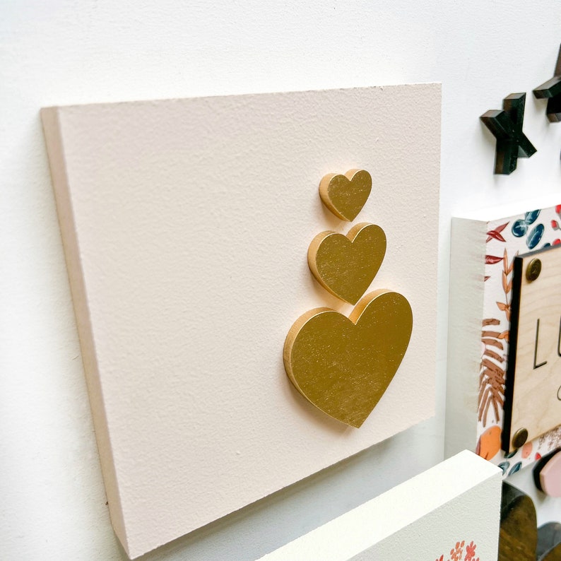 May include: A white canvas with a stack of three gold heart-shaped decorations. The hearts are stacked on top of each other, with the largest heart at the bottom and the smallest heart at the top.