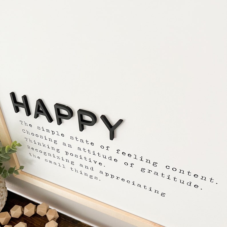 May include: Black wooden letters spelling "HAPPY" on a white background with a definition of happiness in black typewriter font. The definition reads: "The simple state of feeling content. Choosing an attitude of gratitude. Thinking positive. Recognizing and appreciating the small things."