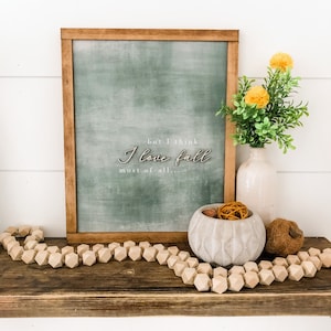 May include: A framed chalkboard sign with the text "but I think I love fall most of all..." in white lettering. The chalkboard is a muted green colour and is framed in a dark wood frame. The sign is sitting on a wooden shelf with a wooden bead garland, a concrete bowl with dried orange peel and a brown ball, and a brown ball of twine.