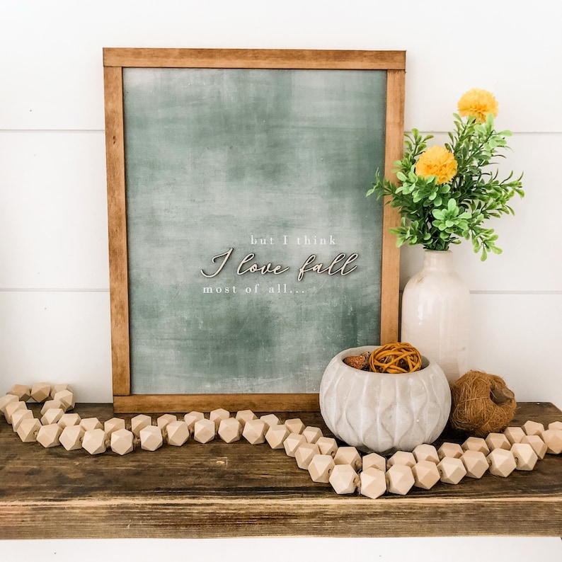 May include: A framed chalkboard sign with the text "but I think I love fall most of all..." in white lettering. The chalkboard is a muted green colour and is framed in a dark wood frame. The sign is sitting on a wooden shelf with a wooden bead garland, a concrete bowl with dried orange peel and a brown ball, and a brown ball of twine.